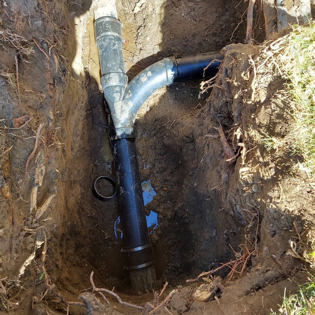 Exposed drainage pipe in a trench with water and soil around it, showing roots nearby.