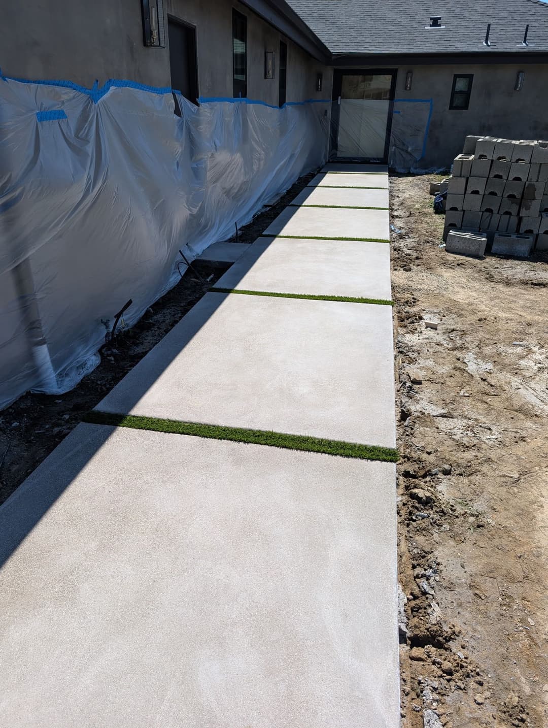Concrete pathway with grass strips, construction site in the background, and protective sheeting.