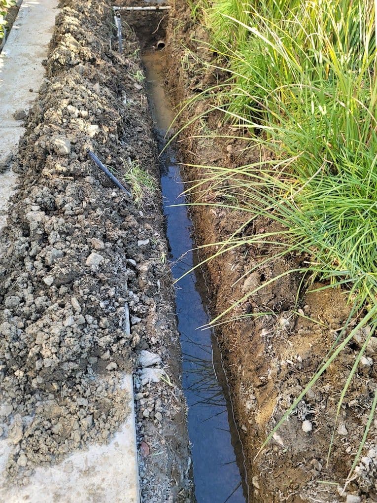 Trench with water in construction site beside grassy area for drainage installation.