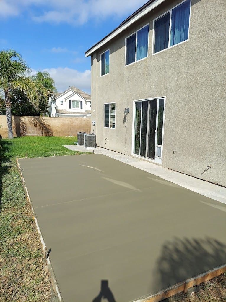 Freshly poured concrete slab in backyard with modern home and palm trees in the background.