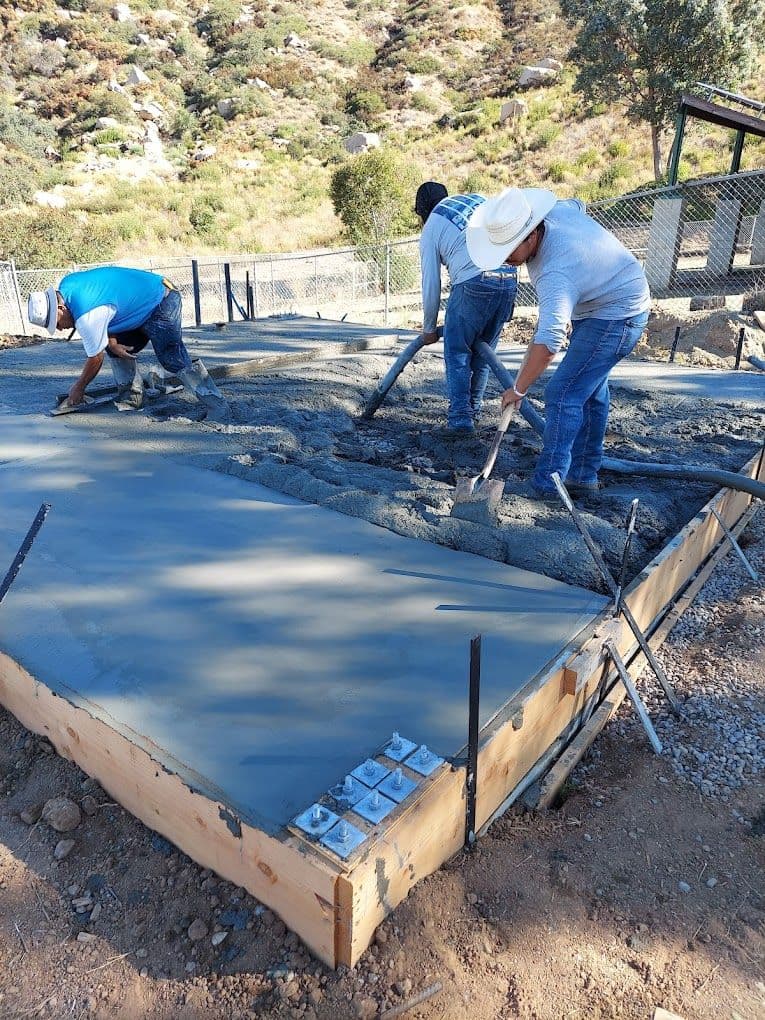 Construction workers pouring and leveling concrete in a rural setting with mountains in the background.
