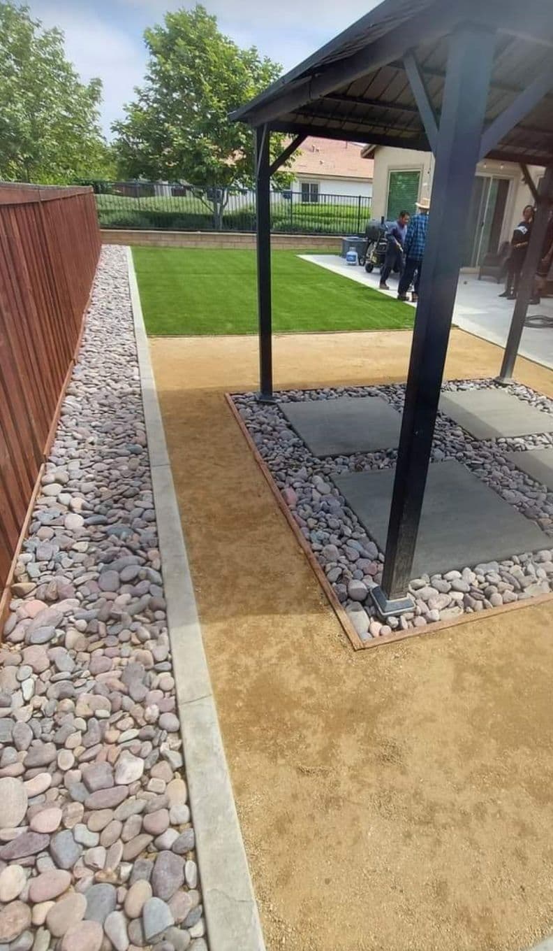Outdoor backyard design featuring gravel pathways, green lawn, and a covered patio area.