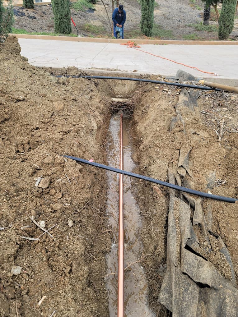 Copper pipe installation in a trench with water flow and a worker in the background.