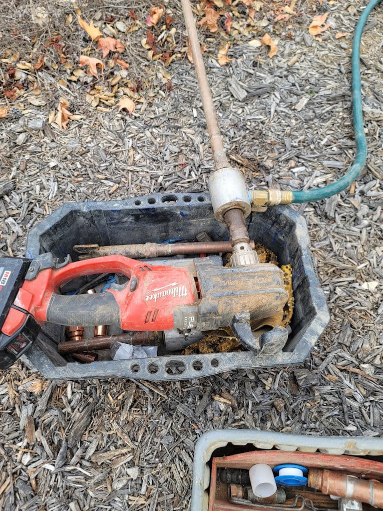 Milwaukee power tool in a plastic bin with plumbing supplies on a mulch surface.