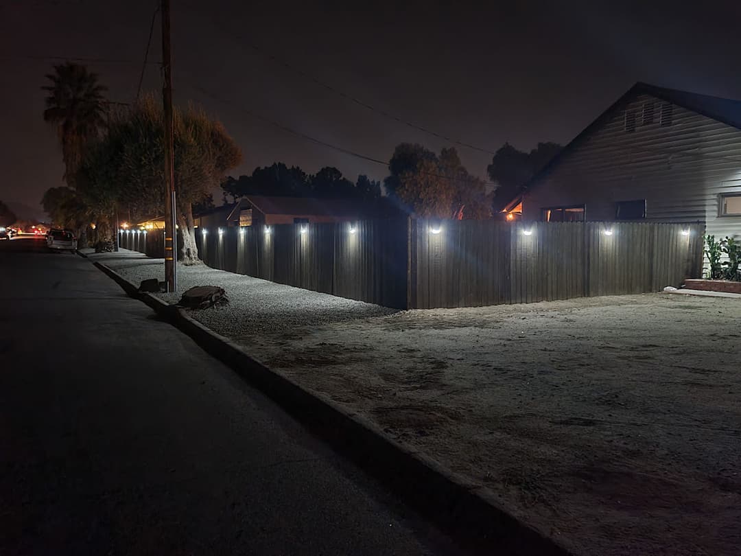 Nighttime street view with illuminated wooden fence and nearby houses along a gravel road.