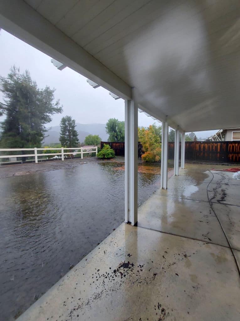 Flooded patio under overhang with rain and distant trees in the background.