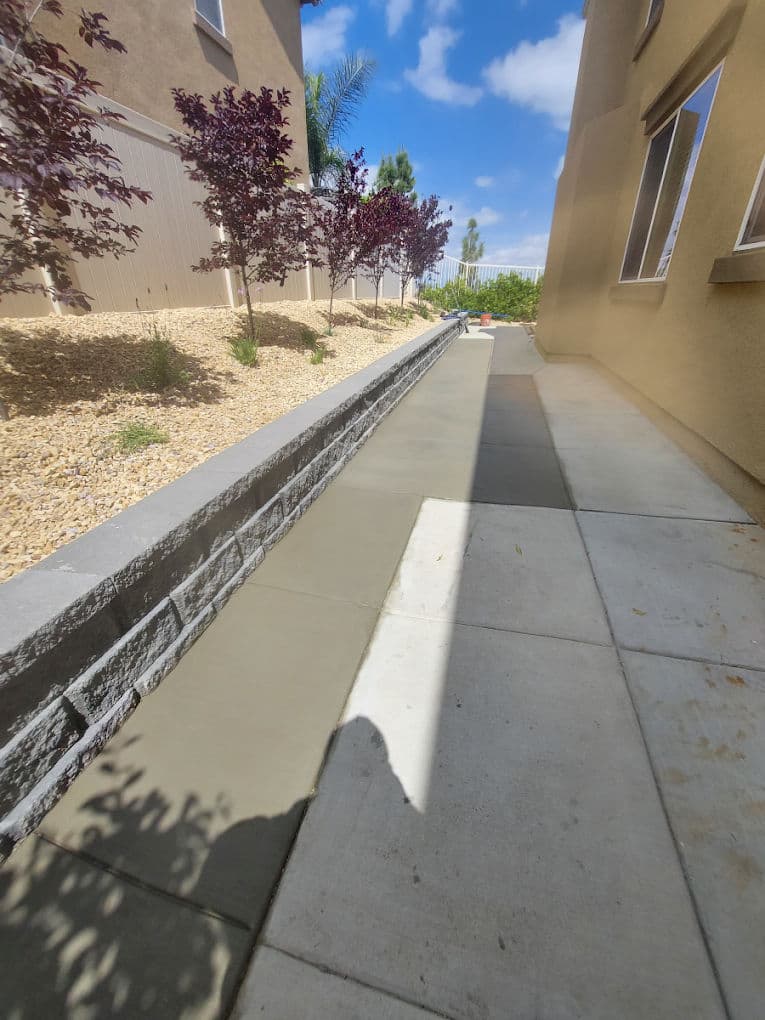 Freshly poured concrete walkway alongside landscaped yard with stone wall and decorative plants.