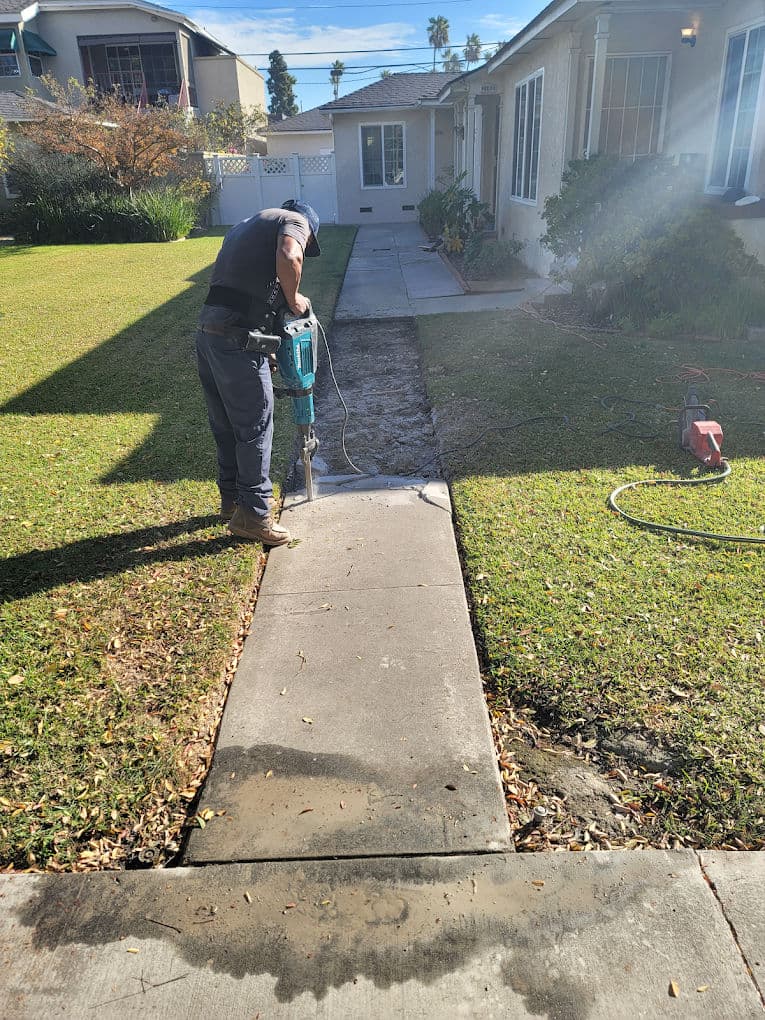 Worker using a jackhammer on a concrete sidewalk in a residential yard, with grass and home in view.