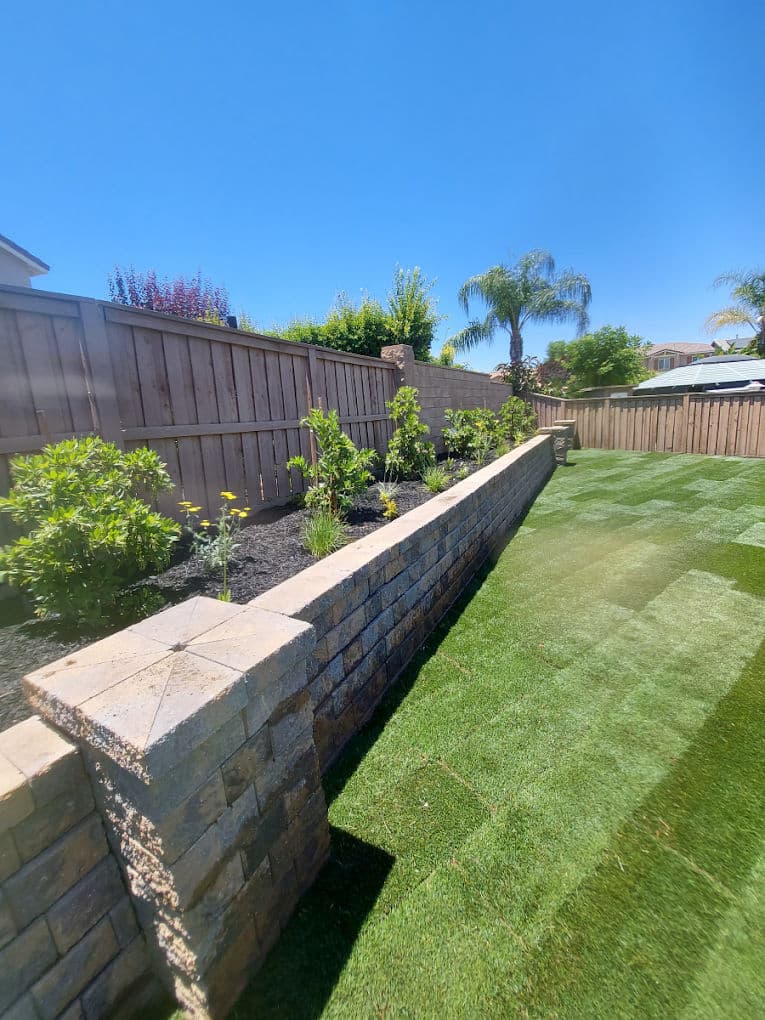 Well-maintained backyard with a stone wall, landscaped plants, and artificial grass under a clear blue sky.