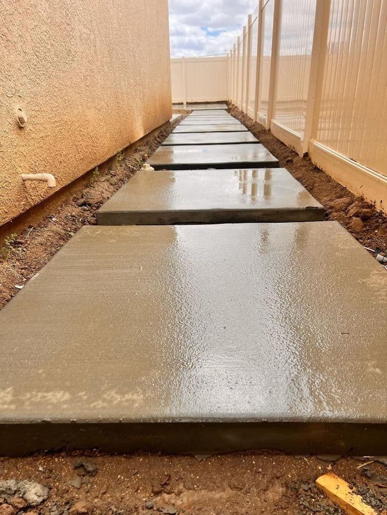 Freshly poured concrete sidewalk between a house and fenced area, showcasing smooth surfaces.
