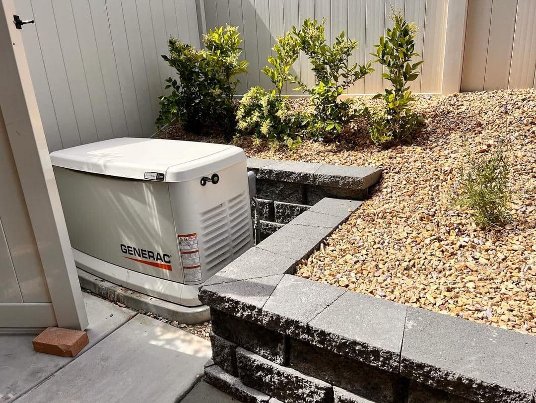 Generac generator in a landscaped yard with stone wall and greenery.