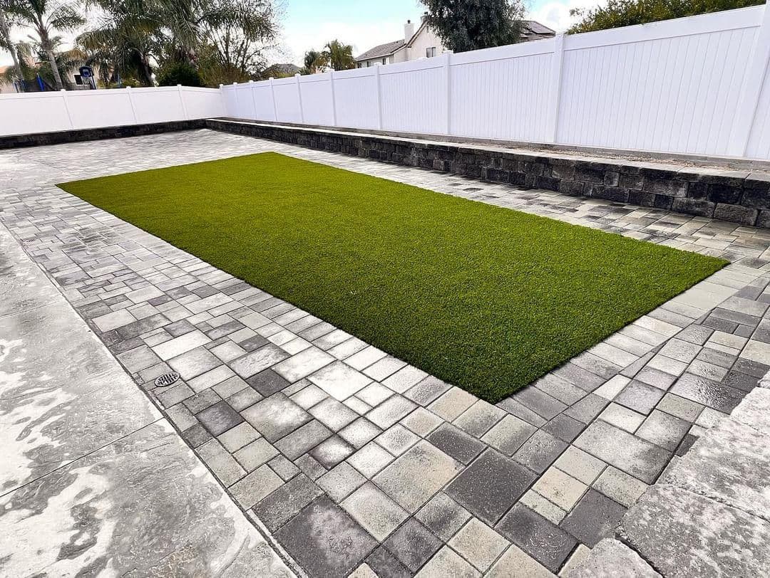 Artificial grass installation on paved patio with white fence and landscaping.