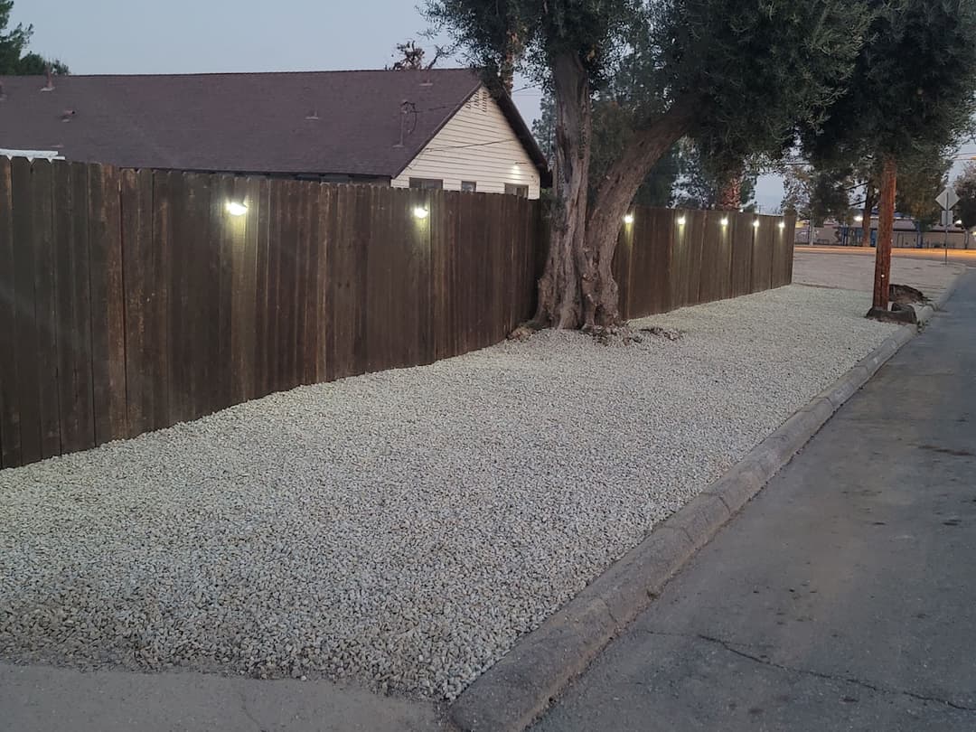 Gravel landscape with low lighting alongside a wooden fence and mature tree at dusk.
