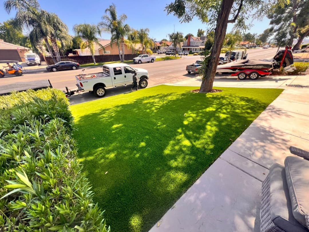 Lush green artificial turf beside a driveway with parked vehicles and palm trees nearby.