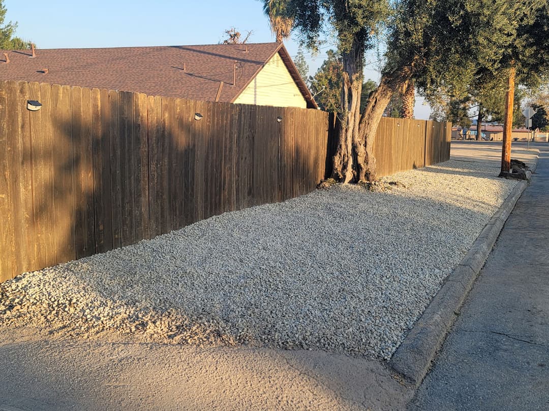 Gravel-covered area beside a wooden fence and tree along a residential street.