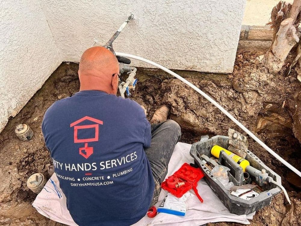Plumber repairing outdoor pipes, showcasing Dirty Hands Services logo and tools.
