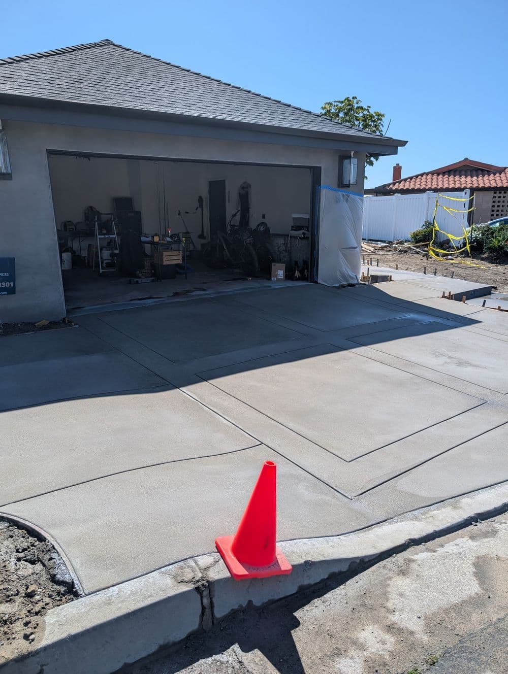 Newly poured concrete driveway in front of a garage with construction tools and safety cone.