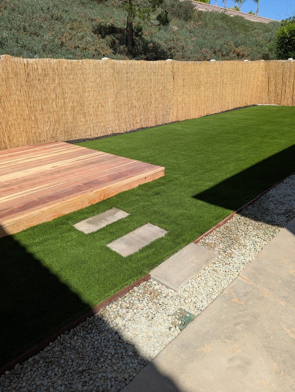 Synthetic grass yard with wooden deck and stepping stones, surrounded by a natural fence.