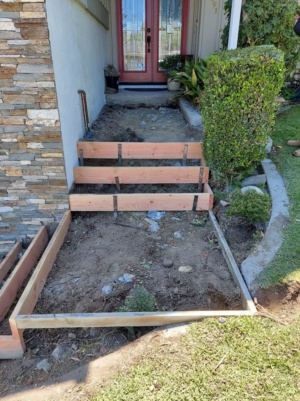 Wooden stairs under construction with stone wall and neatly trimmed bushes in a residential entryway.