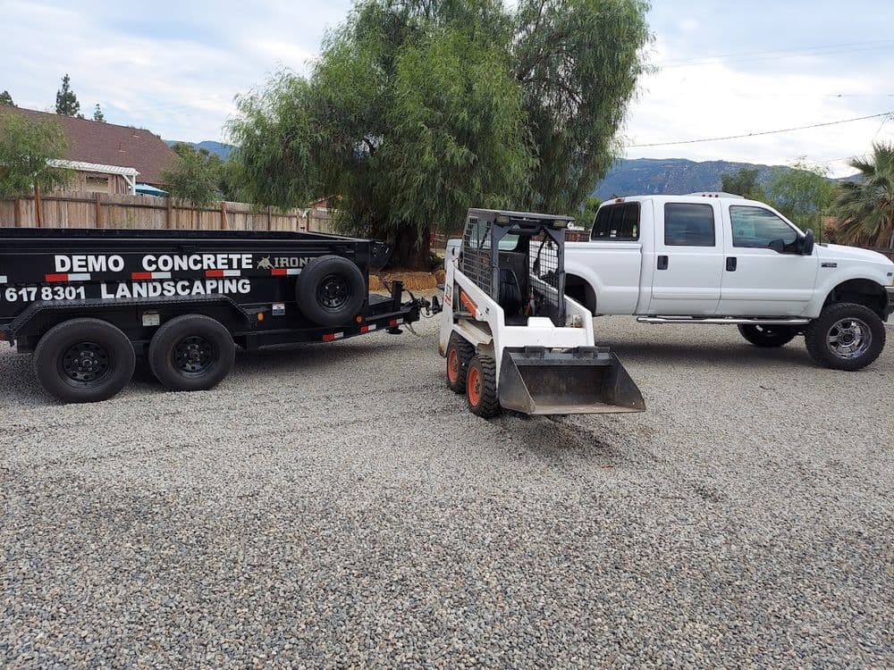 Bobcat skid steer loader and truck with landscaping trailer in gravel area.