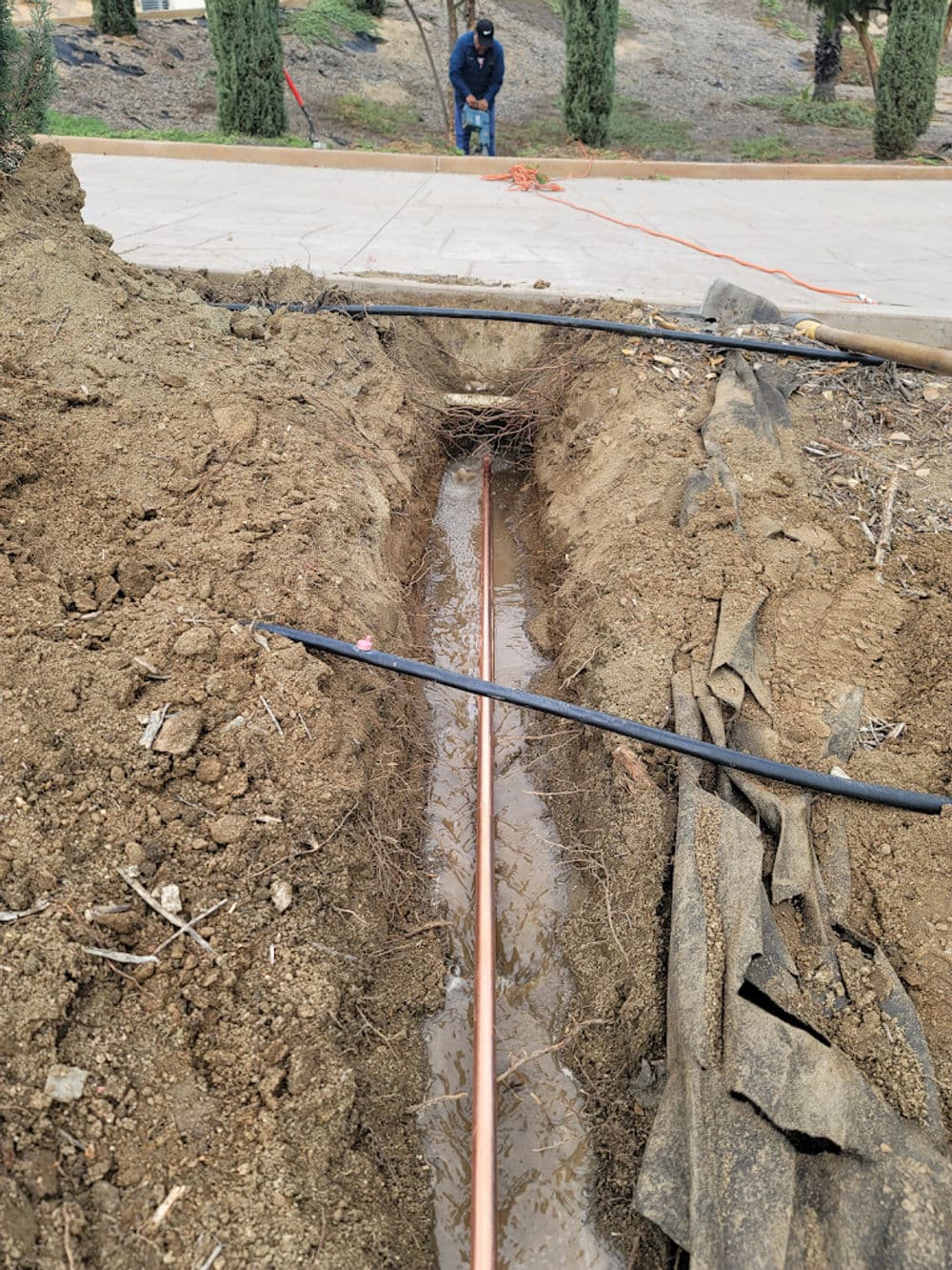 Trench for copper piping installation with a worker and surrounding landscape.