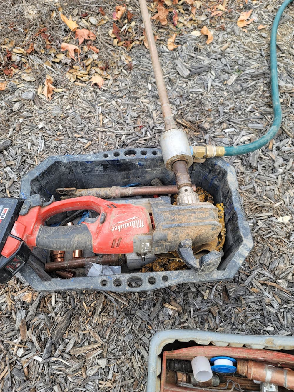Milwaukee power tool and plumbing equipment in a toolbox on a mulch surface.
