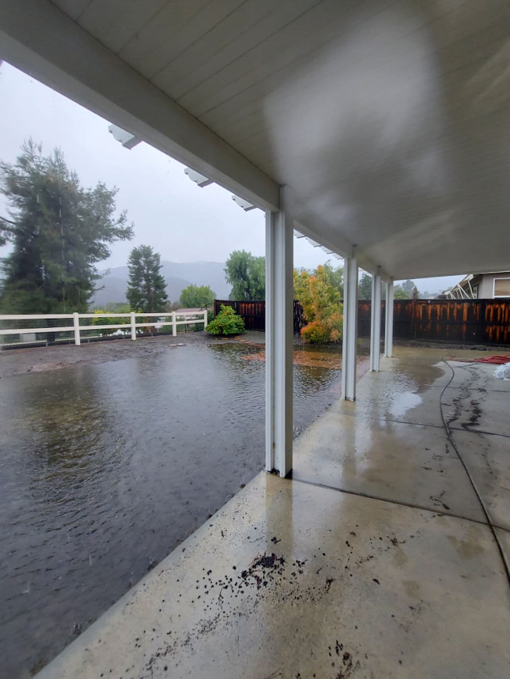 Flooded patio under a covered area, with rain-soaked trees and mountainous background.