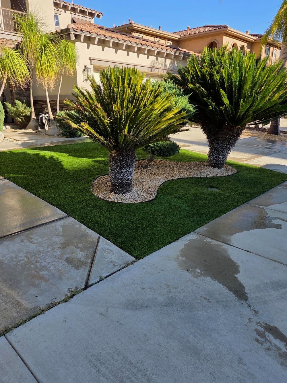 Desert landscape featuring palm trees, decorative gravel, and artificial grass in a sunny setting.
